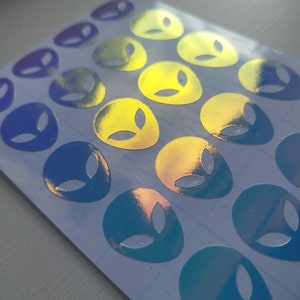 May include: A sheet of holographic alien head stickers in shades of blue, purple, and yellow. The stickers are arranged in rows and are ready to be used for decorating.