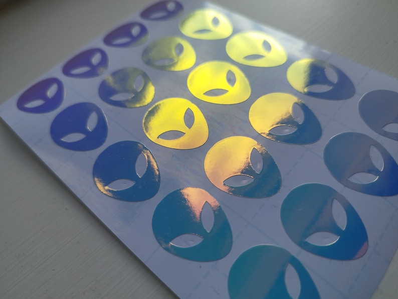 May include: A sheet of holographic alien head stickers in shades of blue, purple, and yellow. The stickers are arranged in rows and are ready to be used for decorating.