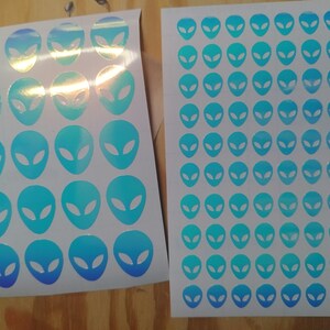 May include: Two sheets of white paper with holographic blue alien head stickers. The stickers are arranged in a grid pattern.