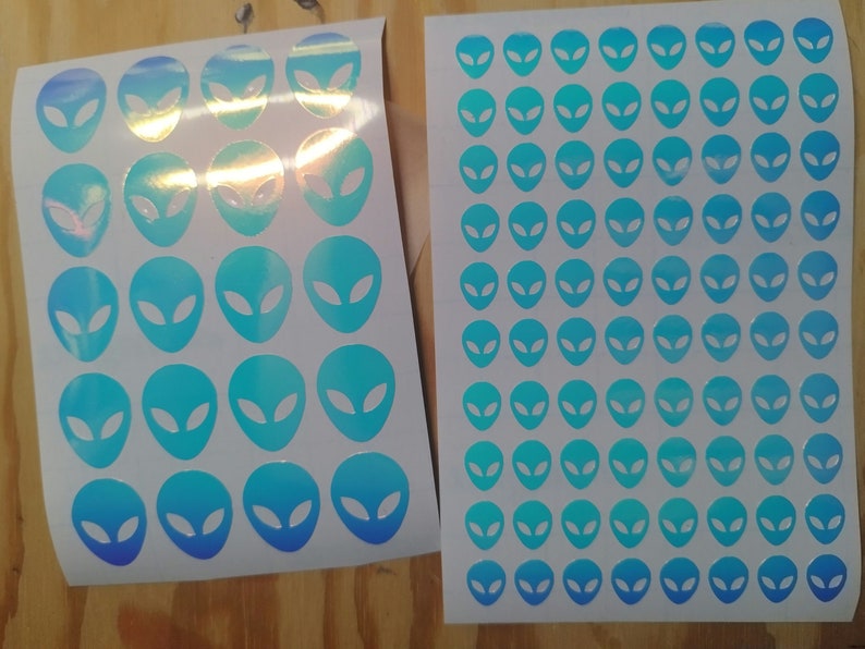 May include: Two sheets of white paper with holographic blue alien head stickers. The stickers are arranged in a grid pattern.