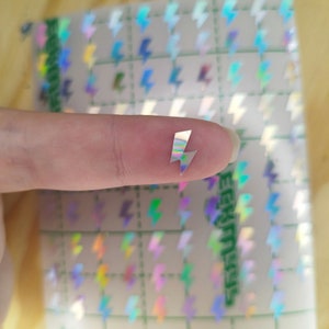 Könnte beinhalten: Ein kleiner, holografischer Aufkleber in Form eines Blitzes auf einem Finger, platziert vor einem größeren Bogen mit mehreren identischen Blitz-Aufklebern, die in einem Raster angeordnet sind, wobei grüne Gitterlinien auf dem Trägerpapier sichtbar sind.