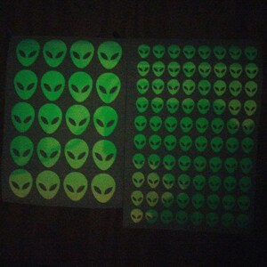 May include: A sheet of glow-in-the-dark stickers featuring alien faces. The stickers are arranged in a grid pattern and are green with a yellow gradient.