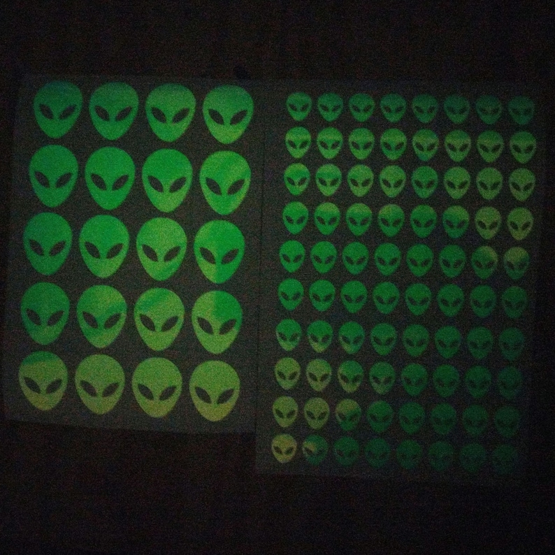 May include: A sheet of glow-in-the-dark stickers featuring alien faces. The stickers are arranged in a grid pattern and are green with a yellow gradient.