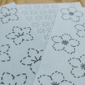 May include: Four white fabric squares with black stitching in a floral pattern. The squares are arranged in a staggered pattern, with the top square partially covering the square below it.