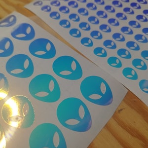 May include: Two sheets of white sticker paper with holographic alien face stickers. The stickers are a gradient of blue and green.