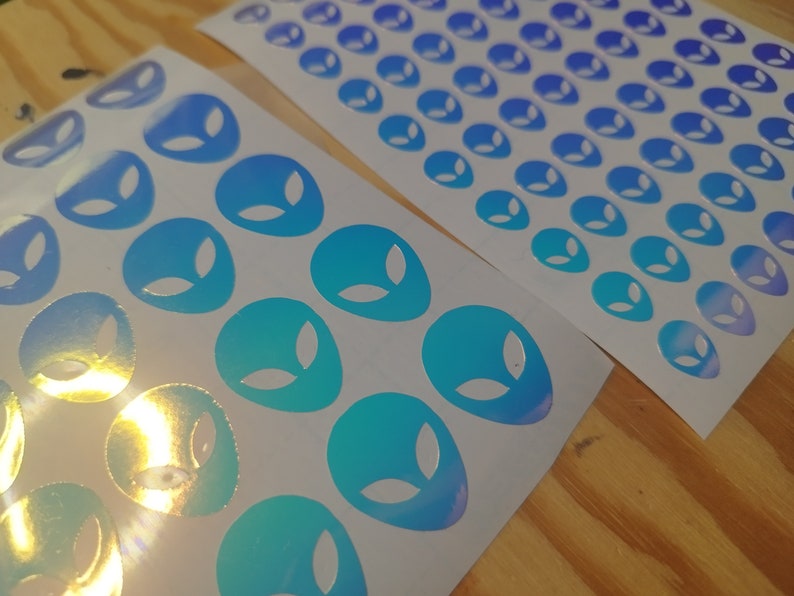 May include: Two sheets of white sticker paper with holographic alien face stickers. The stickers are a gradient of blue and green.