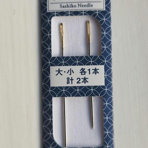 Op de afbeelding: Een verpakking met twee sashiko-naalden, een grote en een kleine, in een blauwe en witte doos met de tekst "Sashiko Needle" en "Olympus".