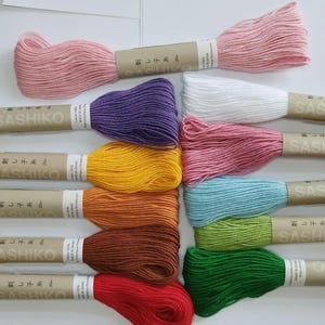 Puede incluir: Una colección de 12 carretes de hilo sashiko en varios colores, incluyendo rosa, morado, amarillo, naranja, marrón, rojo, verde y azul. Cada carrete está etiquetado con el texto japonés "刺し子糸" que se traduce como "hilo sashiko".