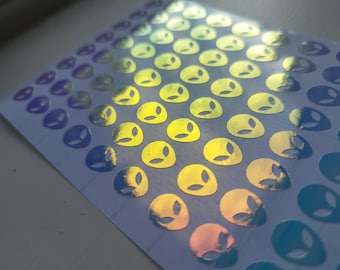 May include: A rectangular sheet of small, circular stickers featuring a die-cut alien head design. The stickers have a reflective, holographic finish that shifts between shades of yellow, blue, and purple depending on the light and angle.