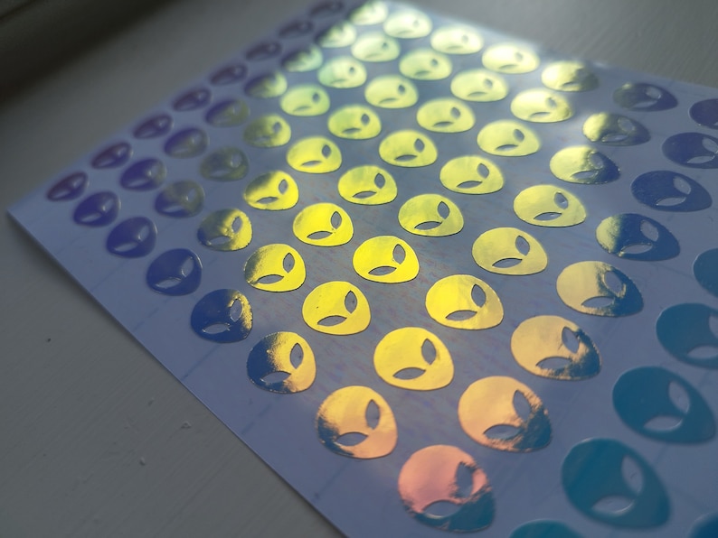 May include: A sheet of holographic stickers featuring alien faces in a gradient of blue, green, and yellow.