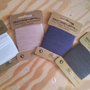 Puede incluir: Cuatro carretes de hilo de algodón para zurcir Scanfil en blanco, rosa, azul marino y gris. Cada carrete está etiquetado con el nombre de la marca y texto en varios idiomas. Los carretes están sobre una superficie de madera marrón claro.