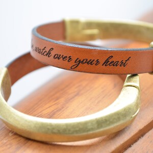 May include: Two brown leather bracelets with gold metal clasps. The top bracelet has the words "watch over your heart" engraved on it.