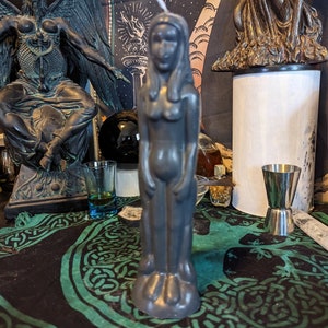 May include: A gray candle shaped like a human figure with long hair. The candle is standing on a black and green patterned surface.