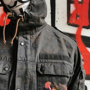 May include: A black hooded jacket with a large pocket on the left side. The pocket has a colorful anarchy symbol painted on it. The person wearing the jacket is wearing a white mask with a black mustache and goatee.
