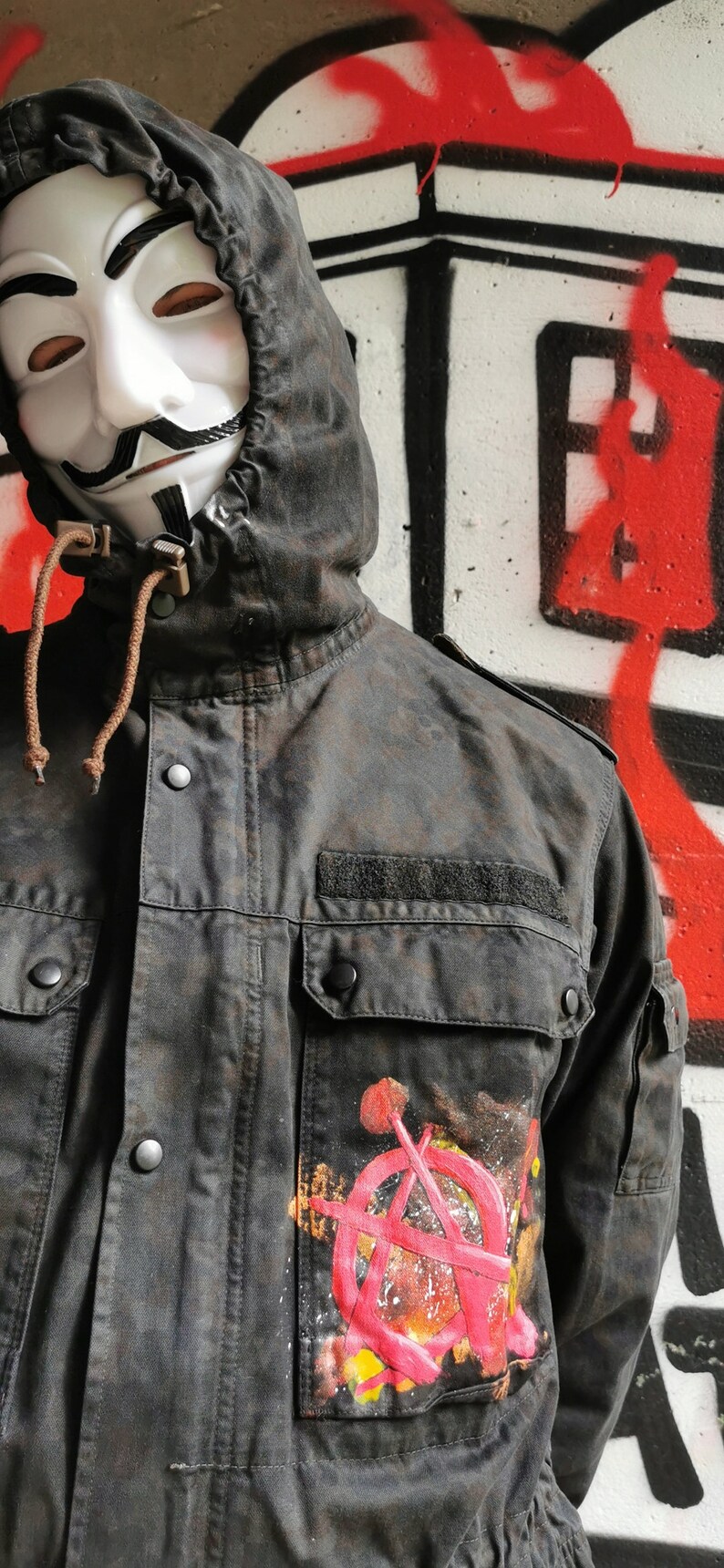 May include: A black hooded jacket with a large pocket on the left side. The pocket has a colorful anarchy symbol painted on it. The person wearing the jacket is wearing a white mask with a black mustache and goatee.