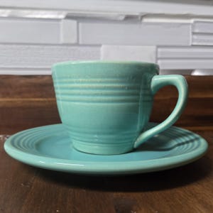 May include: A turquoise ceramic teacup and saucer set. The cup has a rounded shape with a handle and a smooth, glazed finish. The saucer has a slightly scalloped edge and a matching glaze.
