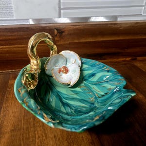May include: A teal ceramic dish with a gold leaf design and a white flower with gold accents. The dish has a handle shaped like a leaf with a gold finish.