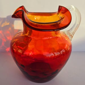 May include: A red and orange glass pitcher with a clear glass handle. The pitcher has a wide mouth and a rounded body.