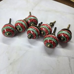 May include: A set of six round cabinet knobs featuring a red and green geometric pattern with white line details. Each knob has a central brass-toned metal post and a matching brass-toned screw base, designed for attachment to furniture or cabinetry.