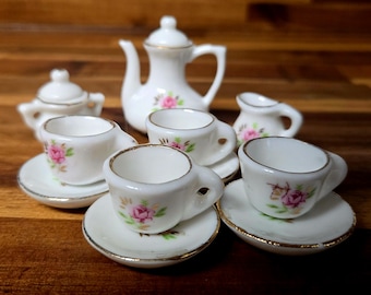 May include: A miniature tea set with a white teapot, creamer, sugar bowl, and four teacups and saucers. The set is decorated with pink roses and gold trim.