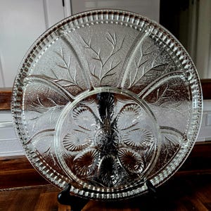 May include: A clear, round glass serving platter with a textured leaf and floral design. The platter is divided into sections and has a decorative beaded edge. It is supported by a black stand.