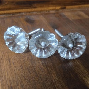 May include: Three clear glass cabinet knobs with a scalloped, flower-like shape and a fluted design. Each knob features a central metal screw post extending from the back, designed for attachment to furniture drawers or cabinet doors.