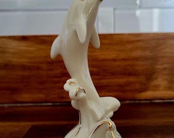 May include: A white porcelain figurine of a dolphin leaping out of the water. The dolphin is painted with gold accents and is set on a base with waves.