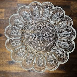 May include: A clear glass egg serving platter with a central circular area and 12 oval-shaped egg compartments. The platter has a textured surface with a scalloped edge, perfect for serving deviled eggs or other appetizers.