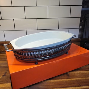 May include: White oval ceramic baking dish with a black metal stand. The stand has a decorative design and four feet. The dish is sitting on a bright orange box.