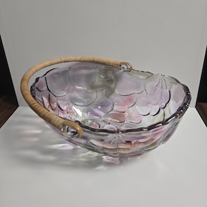 May include: Iridescent glass fruit bowl with a woven handle. The bowl has a grape cluster design and a light purple and pink color. The handle is made of woven tan material. The bowl is oval-shaped and sits on a white surface.