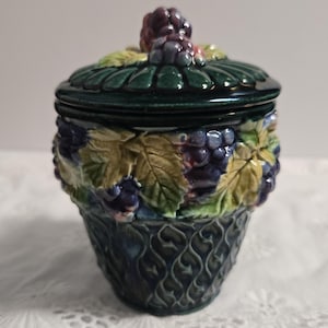 May include: A ceramic jar with a lid, featuring a dark green base and lid, adorned with a grape cluster and leaf design. The jar's body is decorated with a basket weave pattern and grape clusters in shades of purple and green.