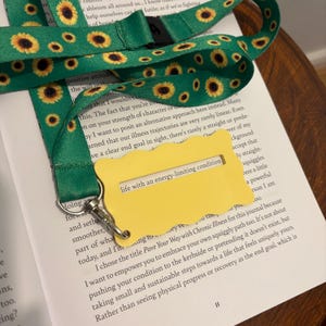 May include: Green lanyard featuring a sunflower design and a yellow tag with the text "life with an energy-limiting condition." The lanyard rests on an open book.