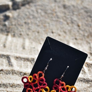 May include: A pair of square-shaped earrings crafted from knotted thread in a gradient of red, orange, and yellow. The earrings feature intricate, looped patterns and are attached to a black rectangular display card with silver-toned metal hooks.
