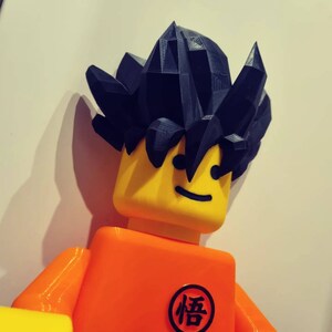 Op de afbeelding: Een geel en oranje Lego-figuur met een zwart stekelig haarstuk. Het figuur heeft een glimlachend gezicht en een zwart kanji-symbool op de borst dat "悟" leest.