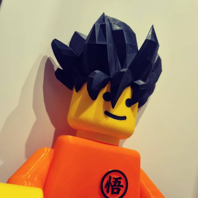 Op de afbeelding: Een geel en oranje Lego-figuur met een zwart stekelig haarstuk. Het figuur heeft een glimlachend gezicht en een zwart kanji-symbool op de borst dat "悟" leest.