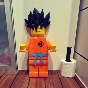 Op de afbeelding: Een groot oranje en geel Lego-figuur van een personage met zwart stekelig haar. Het figuur draagt een blauwe riem en heeft een symbool op de borst met de tekst "悟". Het figuur staat naast een witte toiletrol op een zwarte en bruine houten vloer.