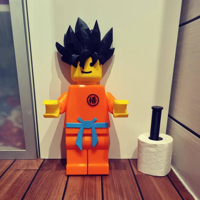 Op de afbeelding: Een groot oranje en geel Lego-figuur van een personage met zwart stekelig haar. Het figuur draagt een blauwe riem en heeft een symbool op de borst met de tekst "悟". Het figuur staat naast een witte toiletrol op een zwarte en bruine houten vloer.