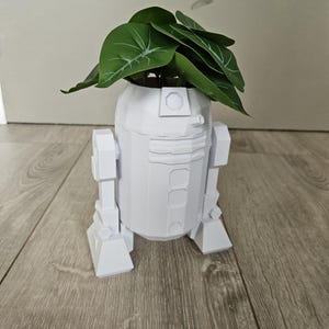 Peut inclure: Un pot de fleurs blanc imprimé en 3D en forme du personnage de Star Wars R2-D2, avec une plante verte à feuilles.