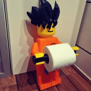 Op de afbeelding: Een 3D-geprinte toiletpapierhouder in de vorm van een Lego-figuur met zwart haar en een geel gezicht. De figuur houdt een rol wit toiletpapier vast.