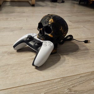 Può includere: Un supporto per console di gioco a forma di teschio nero e oro con un controller di gioco bianco e nero. Il teschio presenta dettagli intricati e un cavo USB si estende dal supporto. Il controller è appoggiato sul supporto.