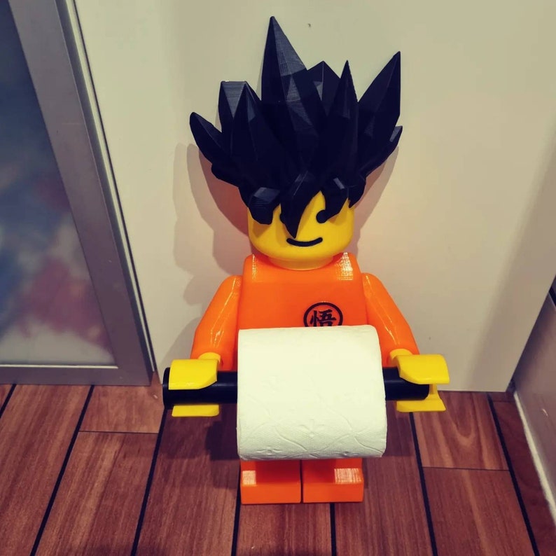 Op de afbeelding: Een zwart en oranje 3D-geprint figuur van een personage met stekelig zwart haar dat een witte rol toiletpapier vasthoudt. Het figuur houdt de rol toiletpapier voor zich vast, en de rol toiletpapier is bevestigd aan een zwart en gele houder.