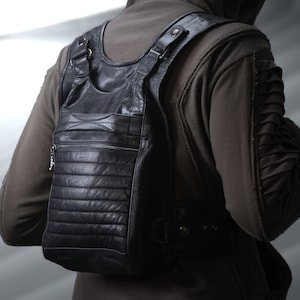 Könnte beinhalten: Schwarzer Lederrucksack mit vertikaler Reißverschlusstasche und horizontalen Steppdetails. Die Tasche hat verstellbare Träger und einen oberen Griff. Die Person trägt eine braune Kapuzenjacke.
