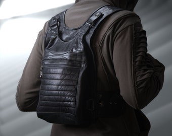 May include: Black leather backpack with a vertical zip pocket and horizontal stitched detailing. The bag has adjustable straps and a top handle. The person is wearing a brown hooded jacket.