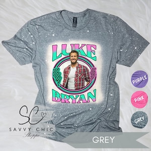 May include: A gray t-shirt featuring a graphic of Luke Bryan in a circle with the text "LUKE BRYAN" in a retro font. The shirt has white paint splatters and color options are shown in the corner.
