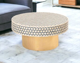 Op de afbeelding: Een ronde salontafel met een wit en zwart geometrisch patroon op de bovenkant en een massief gouden onderstel. De tafel staat op een wit tapijt.