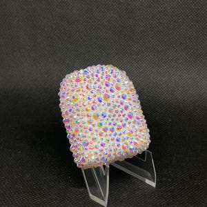 Puede incluir: Un objeto rectangular, posiblemente una funda de teléfono, está cubierto de cristales iridiscentes, exhibido sobre un soporte de acrílico transparente. Los cristales reflejan tonos rosas, amarillos y azules. El fondo es oscuro.