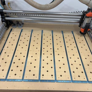 May include: A close-up view of a CNC machine with five wooden panels with evenly spaced holes. The panels are attached to the machine's bed and are ready for processing.