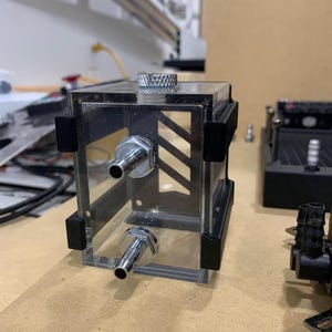 Może przedstawiać: Przezroczysty plastikowy zbiornik z dwoma metalowymi złączami i czarną górą. Zbiornik jest przeznaczony do systemu chłodzenia wodą.