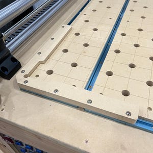 Op de afbeelding: Een close-up van een lichtbruin houten oppervlak met een raster van gaten. Het oppervlak is bevestigd aan een blauwe metalen rail. Het oppervlak maakt deel uit van een houtbewerkingsmachine.