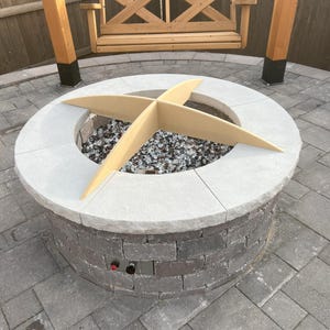 May include: A round, grey brick fire pit with a wooden cross-shaped insert. The fire pit is surrounded by grey paving stones.